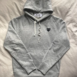 Grå CDG play hoodie - Säljer denna feta hoodien, strl S, från CDG med luva och svart broderat hjärta på vänster sida. Nytt skick, ör av er vid frågor!                                                                                 ⚜️Få ett GRATIS armband vid köp av detta plagg.     Välj mellan ett Anchor eller cuff armband, annars medföljer ett slumpmässigt utav de två⚜️