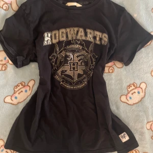 Vintage Hogwarts t-shirt Harry Potter - Svart t-shirt med Hogwarts-tryck i silver och stort emblem på bröstet. Rund halsringning och korta ärmar. Märkt med officiell Harry Potter-logga och tillverkad i mjuk bomull. Snygg till att styla med jeans! För en 2000s outfit Passar storlek XS-S