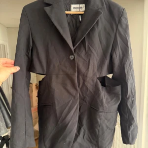 Svart omlottkavaj från Weekday - Solen gör inte bilden rättvis, den är svart och inte missfärga.                                                                              Unik svart kavaj från Weekday i storlek 34 med cool cut-out detalj runt midjan och klassisk krage. Kavajen har en knapp framtill och två fickor. Materialet är troligtvis bomull eller polyester, och modellen är omlott med en edgy twist. Solen gör inte bilden rättvis, den är svart och inte missfärgad 