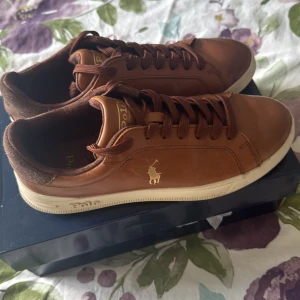 Bruna sneakers från Polo Ralph Lauren - Snygga bruna sneakers från Polo Ralph Lauren med klassisk design och guldfärgad logga på sidan. Skorna har rund tå, snörning och en vit platt sula med Polo-detaljer undertill. Ovandelen är i skinn och mocka, vilket ger en stilren och exklusiv känsla. Kan ändra mig mycket när de kommer till priset vid snabbaffär 