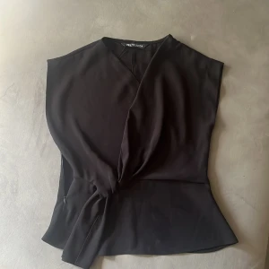 Svart omlottopp från Zara XS - Snygg svart omlottopp från Zara i storlek XS. Toppen har en v-ringad hals och knyt i midjan som ger en cool siluett. Ärmlös design och tillverkad i mjukt polyester-material. Perfekt för dig som gillar stilrena och trendiga plagg.