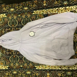 Ljuslila hoodie med broderad logga - Säljer en ljuslila hoodie med broderad logga på bröstet. Tröjan har huva med snörning och en stor magficka framtill. Perfekt för dig som gillar en chill och avslappnad stil. Storlek S/M