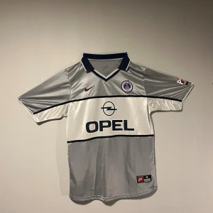 PSG grå retro fotbollströja - Snygg PSG fotbollströja i silver med svarta detaljer och Opel-logga på bröstet. Tröjan har klubbmärke, klassisk krage och är tillverkad i glansigt funktionsmaterial. Perfekt för dig som gillar retrostil och samlar på fotbollströjor.
