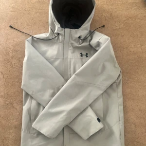 Ljusgrå vindjacka från Under Armour - Snygg ljusgrå vindjacka från Under Armour med huva och justerbara snören. Jackan har en clean look med svart logga på bröstet och dragkedja framtill. Tillverkad i slitstark polyester och har en loose passform, perfekt för dig som gillar sportig stil.