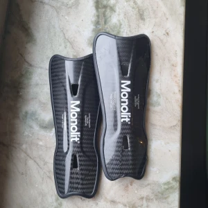 Monolit benskydd i kolfiber - Säljer ett par svarta benskydd från Monolit i kolfiber med snyggt mönster och ergonomisk form. De är lätta och smidiga, perfekta för fotbollsträning eller match. Kommer med en praktisk svart förvaringsväska med dragkedja.