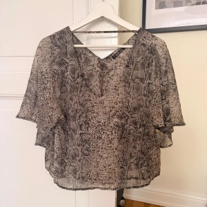 Ormmönstrad blus från LUC.CE - Säljer en luftig och transparent blus från LUC.CE i storlek M. Blusen har ett coolt ormmönster i beige och svart, v-ringning och korta, vida ärmar. Perfekt för dig som vill sticka ut med en trendig och lätt topp.