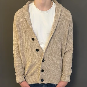 Beige stickad kofta med knappar - Mysig beige stickad kofta med grov struktur och stora svarta knappar framtill. Koftan har en avslappnad passform, ribbade muddar och en bred krage som ger extra chill vibe. Perfekt att slänga på över en t-shirt när det är lite kyligt ute.