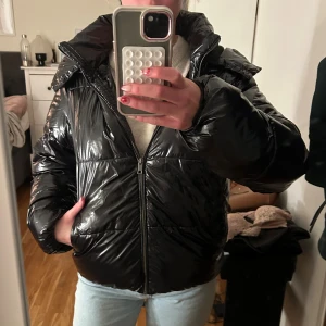 Svart pufferjacka från Nelly med huva - Säljer en svart, glansig pufferjacka från Nelly i storlek M. Jackan har en stor, mysig huva och dragkedja framtill. Den är quiltad och har en loose passform som ger en cool och avslappnad vibe. Perfekt för kalla dagar när du vill vara både varm och snygg.