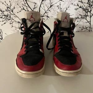 Nike Air Jordan 1 Mid röd/svart/vit 39 - Säljer ett par Nike Air Jordan 1 Mid i klassisk röd, svart och vit färg. Skorna har svart snörning, vit swoosh och detaljer samt hög modell. Tillverkade i skinn och syntet, med rund tå och platt sula. Perfekta för dig som gillar streetstyle och sneakers med ikonisk design.