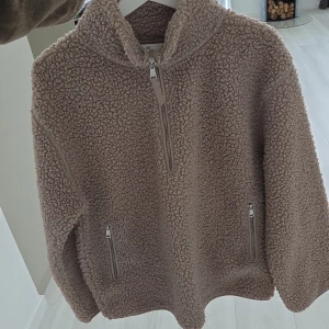 Beige/brun teddy tröja/jacka från H&M - Mysig beige teddyfleece-tröja från H&M med halv dragkedja framtill och två sidofickor med dragkedja. Tröjan har hög krage och lång ärm, perfekt för kyliga dagar. Oversized och fluffig känsla som ger en avslappnad look.Endast använd en gång så den är som ny💕