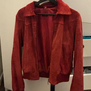 Röd mocka jacka - Snygg röd bomberjacka från Lovit i mockaliknande material. Jackan har klassisk krage, dragkedja framtill och ribbade muddar vid ärmslut och midja. Stilsäkra sömmar och två fickor framtill ger en cool retrovibe. Perfekt för dig som vill sticka ut.