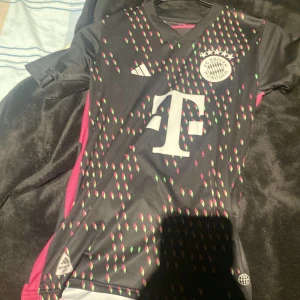 FC Bayern München Musiala tröja - Säljer en FC Bayern München fotbollströja med Musiala och nummer 42 på ryggen. Tröjan är svart med rosa och gröna geometriska mönster, Adidas-logga och klubbmärke på bröstet. Kortärmad modell i lätt och ventilerande polyester, med rosa detaljer på sidorna och axlarna.