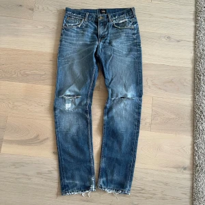 Blå slitna jeans från Lee - Säljer ett par blå jeans från Lee med slitningar och hål på knäna samt fransiga benslut. Jeansen har klassisk femficksdesign, normal passform och är tillverkade i jeansmaterial. Perfekta för en avslappnad och cool stil. Står att det är strl 27/33 men stämmer absolut inte, sitter mer som 30/32. 