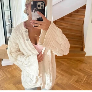 Vit kabelstickad kofta med knappar - Supermysig vit kabelstickad kofta med stora, ribbade ärmslut och knappar framtill. Koftan har en loose passform och är perfekt att slänga över en t-shirt eller linne. Tjockt stickat material som känns både chill och trendigt.