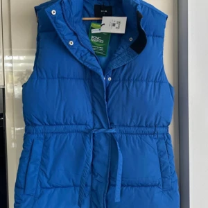 Blå dunväst med knyt från Bik Bok - Snygg blå dunväst från Bik Bok med hög krage, dragkedja och tryckknappar framtill. Västen har praktiska fickor och ett knytband i midjan för att justera passformen. Perfekt för lager-på-lager och chill stil under kyliga dagar.