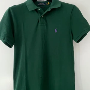 Grön pikétröja Polo Ralph Lauren - Mörkgrön pikétröja från Polo Ralph Lauren med lila broderad logga på bröstet. Tröjan har klassisk krage, korta ärmar och knappar vid halsen. Modellen är custom slim fit och tillverkad i mjuk bomull. Endast använd två gånger så i väldigt fint skick.  