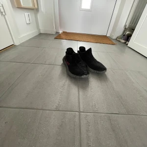 Adidas Yeezy Boost 350 V2 Black/Red - Säljer ett par Adidas Yeezy Boost 350 V2 i svart med röda SPLY-350-detaljer på sidan. Skorna har en stickad ovandel och en gråsvart, räfflad sula med boost-dämpning. Perfekta för dig som gillar streetwear och vill sticka ut med en ikonisk sneaker. Ingen sula i