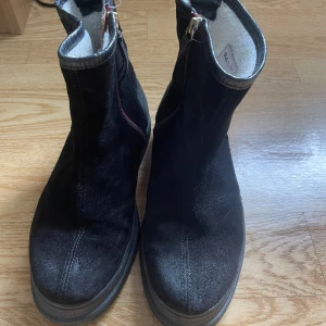 Svarta mockaboots från Gant - Svarta boots från Gant i mjuk mocka med dragkedja framtill och detaljer i skinn. Insidan är fodrad med ull för extra värme. Snygg, stilren design med rund tå och platt sula, perfekta för kyligare dagar.