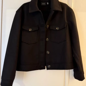 Svart jacka från Vero Moda - Stilren svart jacka från Vero Moda i storlek M. Jackan har klassisk krage, stora bröstfickor med knappar och raka, långa ärmar. Materialet är mjukt och sköntt. Perfekt för lager-på-lager och enkel att matcha.