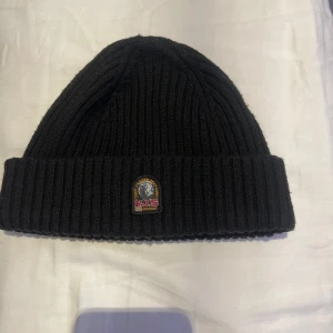  Parajumpers mössa - Svart ribbstickad mössa från Parajumpers med uppvikt kant och klassisk PJS-logga framtill. Tillverkad i mjukt material som håller dig varm under kalla dagar. Perfekt accessoar för en stilren och sportig look.