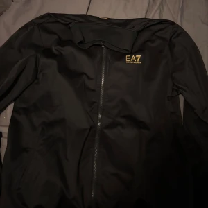 Svart Zip  från EA7 Emporio Armani Storlek L Passar M - Svart Zip h från EA7 Emporio Armani med guldfärgad logga på bröstet och dekorativa guldstreck på ryggen. Zipen har hel dragkedja framtill, hög krage och diskreta fickor. Snygg och stilren design som passar perfekt till streetwear.