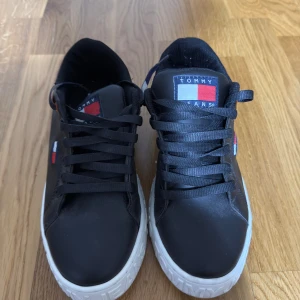 Svarta sneakers Tommy Jeans - Snygga svarta sneakers från Tommy Jeans med vit sula och klassisk logga på sidan och plösen. Skorna har svarta snören, diskreta röda detaljer och är tillverkade i syntetmaterial. Perfekta för dig som gillar stilrena och sportiga skor med en modern touch.