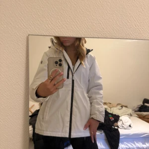 Vit vindjacka från Five Seasons - Säljer en vit vindjacka från Five Seasons med svart dragkedja och huva. Jackan har flera fickor med dragkedja och en sportig look. Perfekt för blåsiga dagar och enkel att matcha med olika outfits. Finns tecken på användning men inget man märker vid första anblick. Ny pris ligger runt 1200kr
