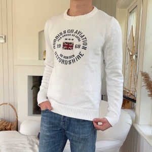 Vit stickad tröja från Morris Gents - Vit stickad tröja från Morris Gents med tryck och brittisk flagga på bröstet. Rund halsringning och ribbade muddar vid ärmslut och nederkant. Perfekt för dig som gillar klassisk och stilren look.