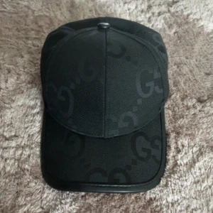 Svart keps  med GG-mönster - Snygg svart keps med diskret GG-logomönster över hela kepsen. Klassisk form med böjd skärm och justerbar rem baktill i svart och guld. Tillverkad i slitstark bomull med exklusiv känsla, perfekt för dig som gillar designeraccessoarer.