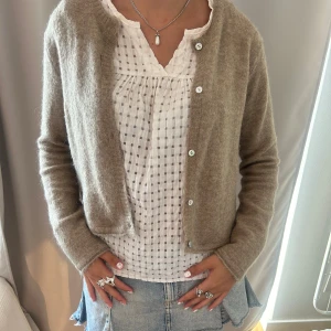 Beige stickad kofta med knappar - Supermysig beige stickad kofta med rund hals och knäppning framtill. Koftan är croppad och har långa ärmar, perfekt att slänga över en topp eller klänning. Enkel och stilren design som passar till jeans eller kjol.
