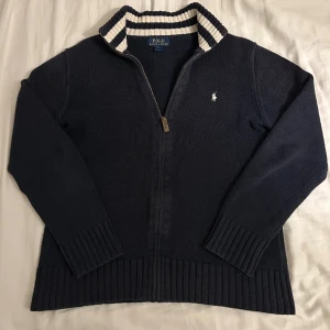 Mörkblå kofta från Polo Ralph Lauren - Mörkblå helzip från Ralph Lauren. Riktigt snygg med de vita ränderna på kragen och har en bra passform. Tröjan är varm och är i storlek L 14-16 år så tröjan passar alla inom storleken S. Kontakta vid intresse!
