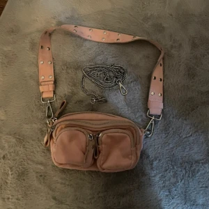 Rosa axelväska från Gina Tricot - Snygg rosa axelväska från Gina Tricot med bred axelrem dekorerad med silvriga nitar . Väskan har även en avtagbar kedja som gör att man kan bestämma själv vilken man vill ha på,  och är tillverkad i mjukt syntetmaterial.