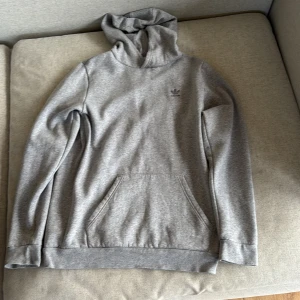 Grå hoodie  - Säljer en stilren grå hoodie från Adidas med broderad logga på bröstet. Tröjan har huva, magficka och ribbade muddar. Passar dig runt 175