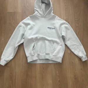 Grå Essentials Fear of God hoodie - Säljer en ljusgrå hoodie från Essentials Fear of God med svart logga på bröstet och stor text på ryggen. Tröjan har huva, känguruficka och är långärmad. Oversized passform och tillverkad i mjuk bomullsblandning. Perfekt för en chill och trendig look.