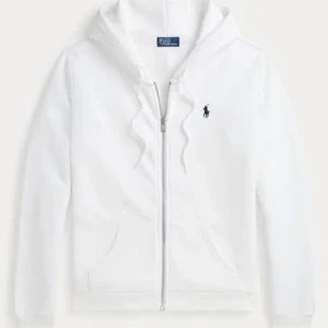 Vit hoodie från Polo Ralph Lauren - Vit hoodie från Polo Ralph Lauren med dragkedja framtill, huva med snörning och broderad blå logga på bröstet. Klassisk passform och mjukt material, perfekt för en chill look.