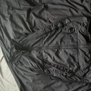 Svart dunväst från C.P. Company - Kan fås mer bilder för bevis på att den är äkta. Svart dunväst från C.P. Company med Primaloft-foder. Västen har dragkedja framtill, två snedställda fickor med knappstängning och den ikoniska linsen på bröstet. Lätt och smidig design, perfekt för lager-på-lager. Snygg och funktionell för kyligare kvällar eller bara när som. Helt ny använd 3 gånger då den är förliten på mig köpte fel storlek 