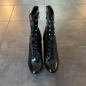 Svarta platåstövletter med snörning - Säljer ett par svarta, glansiga stövletter med snörning och dragkedja på sidan. Skorna har hög platå och smal, genomskinlig klack i plast. Materialet är lackliknande syntet och de har rund tå. Perfekta för dig som vill sticka ut med en edgy look.