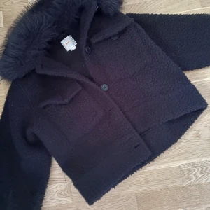 Svart teddyjacka med fuskpäls - Mysig svart teddyjacka från Gina tricot med fluffig fuskpäls runt kragen. Jackan har två stora fickor framtill, knäpps med knappar och har en boxig, lite croppad passform. Perfekt för kalla dagar när du vill vara både varm och snygg.