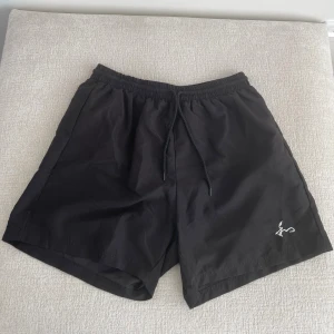 Svarta shorts från Represent - Svarta shorts från Represent med enkel design och vit broderad logga på ena benet. De har elastisk midja med snörning för skön passform. Perfekta för en avslappnad stil under varma dagar.