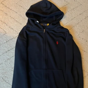 Svart hoodie från Polo Ralph Lauren - Mörkblå hoodie från Polo Ralph Lauren med dragkedja och huva. Klassisk röd broderad logga på bröstet och känguruficka framtill. Tillverkad i mjuk bomullsmix, perfekt för chill dagar. Snygg och enkel att matcha med jeans eller joggers. Använd några fåstaka gånger med ser ut som helt ny. Pytteliten fläkt på tröjan som borde gå bort med tvätt men annars inte syns. Fraktar inom 1-2 dagar! 