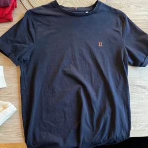 Svart t-shirt från Les Deux - Svart t-shirt från Les Deux med enkel design och liten broderad logga i orange på bröstet. Klassisk rund halsringning och korta ärmar. Tillverkad i mjuk bomull för en skön känsla. Perfekt för en clean och stilren look.