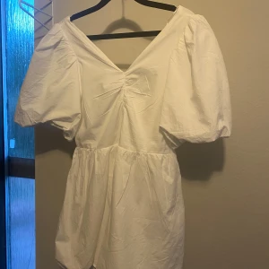 Vit kort puffärmad klänning Gina Tricot - Superfin vit kort klänning från Gina Tricot med puffärmar, v-ringning och söt rosettdetalj i ryggen. Klänningen har en luftig passform och är gjord i bomull, perfekt för varma dagar. Enkel men ändå trendig design som sticker ut med sina detaljer. Endast använd en gång😊🥰🤗