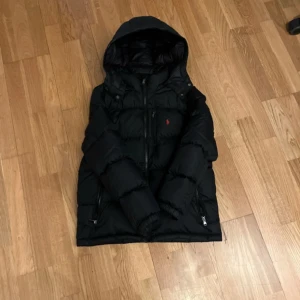 Svart dunjacka Polo Ralph Lauren - Svart dunjacka från Polo Ralph Lauren med huva och dragkedja framtill. Jackan har det klassiska röda Polo-logot broderat på bröstet och två fickor med dragkedja. Perfekt för kalla dagar och riktigt snygg streetstil. Väldigt bra skick hör av er för frågor.