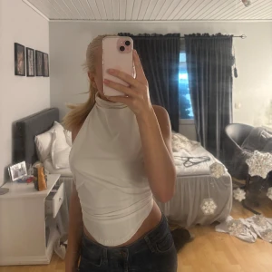 Vit halterneck topp med öppen rygg - Säljer en vit halterneck topp med hög krage och öppen rygg. Toppen knyts i ryggen med tunna band och har en asymmetrisk nederkant framtill. Perfekt för dig som vill ha en stilren och trendig look.