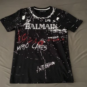 Svart Balmain t-shirt med graffiti-tryck - Cool svart t-shirt från Balmain med unikt graffiti-inspirerat tryck i vitt och rött både fram och bak. T-shirten har korta ärmar och rund hals. Materialet är mjuk bomull som känns skön mot huden. Perfekt för dig som vill sticka ut med en edgy streetstyle.