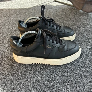 Svarta sneakers från Axel Arigato - Snygga svarta sneakers från Axel Arigato med vit, chunky sula och klassisk snörning. Skorna är i skinn och har diskret guldtext på sidan. Perfekta för dig som gillar stilrena och trendiga sneakers med en modern vibe.