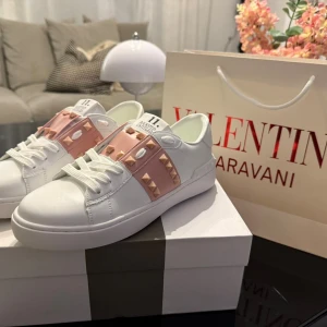 Valentino Garavani Rockstud sneakers - Snygga vita Valentino Garavani Rockstud sneakers med rosa band och ikoniska nitar. Skorna har rund tå, snörning och platt sula. Tillverkade i skinn med exklusiv känsla och detaljer som sticker ut. Perfekta för dig som vill ha något extra till din outfit.