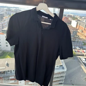 Svart pikétröja från Bläck, storlek M - Klassisk svart pikétröja från Bläck i storlek M. Tröjan har korta ärmar, krage och knapplös öppning framtill. Perfekt för en clean och stilren look. Materialet är mjukt och bekvämt, passar till många olika outfits.