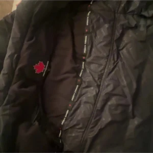 Svart pufferjacka från Zavetti Canada - Säljer en svart pufferjacka från Zavetti Canada med huva och tydlig logga på ärmen. Jackan har quiltad design, dragkedja framtill och är perfekt för kalla dagar. Fodret är tjockt och jackan har en modern, lite oversized look.