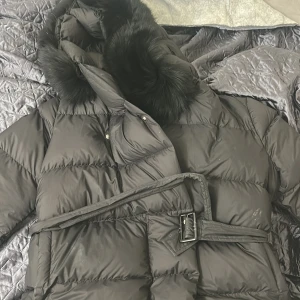Svart pufferjacka från FUR Stockholm - Snygg svart pufferjacka från FUR Stockholm med stor fluffig päls /äkta på huvan och bälte i midjan. Jackan har quiltad design och stängs med både knappar och bälte. Perfekt för kalla dagar när du vill vara både varm och stilren.